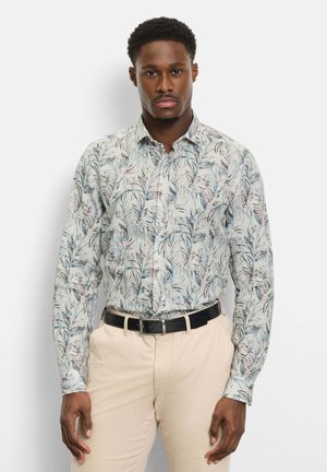 Mann trägt ein helles, langärmeliges Hemd mit floralem Muster, in beige Hosen mit schwarzem Gürtel gesteckt, steht vor einem schlichten weißen Hintergrund.