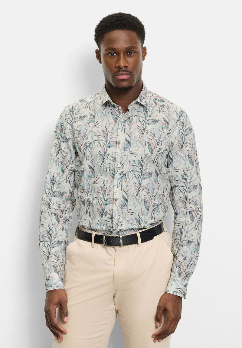 Homme portant une chemise à manches longues à motif floral clair rentrée dans un pantalon beige avec une ceinture noire, debout devant un fond blanc uni.