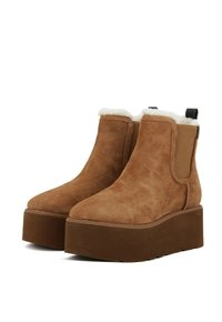 Stivali ankle in suede marrone con un morbido rivestimento bianco, suole piatte spesse marroni, pannelli elastici laterali e una linguetta sul retro.