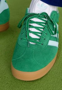 Tenis de camurça verde com detalhes brancos e sola de borracha texturizada. Apresenta atacadores verdes com um padrão riscado. Detalhes de costura visíveis.