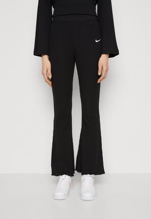 Personne portant un pantalon évasé côtelé noir avec un logo blanc Nike et des baskets blanches, debout devant un fond clair uni.