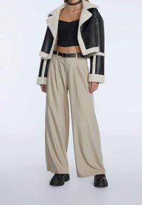 Chaqueta corta de cuero negra con ribete de piel sintética blanca, combinada con pantalones de pierna ancha beige y zapatos de plataforma negros.