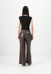 Donna con capelli scuri e lisci che indossa un top senza maniche nero, pantaloni in pelle marroni a gamba larga e tacchi neri, rivolta verso un muro bianco.