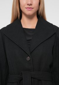 Melnas vilnas mēteles ar plašu apkakli, vienu pogu un sasietu vidukli. Raksturīga gluda virsma un pielāgota forma.