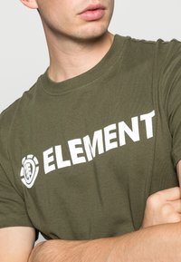 Olivově zelené bavlněné tričko s bílým logem a emblémem "ELEMENT" na levé straně. Krátké rukávy a kulatý výstřih.
