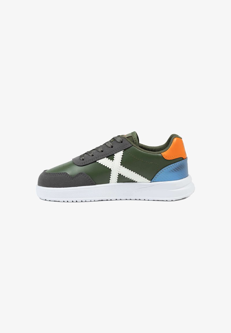 Zapatilla en verde y gris, con costuras blancas, acentos en naranja y azul, parte delantera con cordones y suela texturizada. Materiales lisos y de ante.
