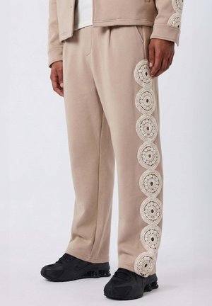 Pantalones beige con siete parches redondos de crochet que recorren el lado exterior de la pierna derecha, combinados con zapatillas negras.