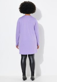 Lila langärmliges Kleid mit abgerundetem Saum und glatter Textur, kombiniert mit glänzenden schwarzen Leggings und mit Nieten besetzten schwarzen Knöchelstiefeln.