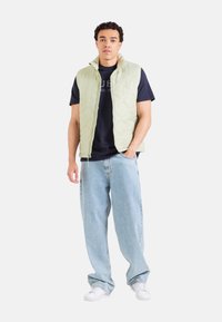 Chaleco acolchado verde claro sobre una camiseta azul oscura, combinado con jeans sueltos de color azul claro y zapatillas blancas. El chaleco tiene un cuello alto.