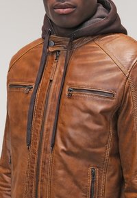 Veste en cuir marron avec finition texturée, dotée de deux poches poitrine zippées et d'une capuche à cordon de serrage foncé. Coupe ajustée et détails cousus.