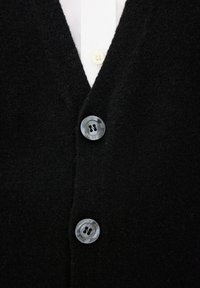 Zwarte cardigan van zachte wol met een V-hals ontwerp, voorzien van twee grijze knopen met vier gaten aan de voorkant, over een wit shirt.
