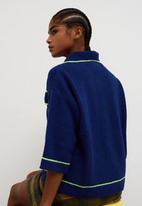 Pulover tricotat supradimensionat bleumarin cu mâneci scurte, având cusături accent color verde de-a lungul gulerului și tivului. Material moale, texturat.