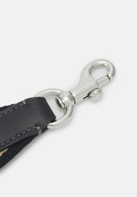 Mousqueton pivotant en métal argenté fixé à une sangle en cuir noir et tissu sur un fond clair uni.