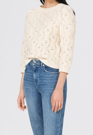 Femme portant un pull en maille crème avec un motif de feuilles et un jean bleu taille haute, debout devant un fond uni clair.