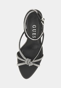 Svart högklackad sandal med spetsig tå, som har korsande rhinestone-remmar och en slät texturerad innersula med "GUESS"-logotyp.