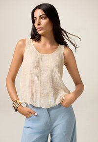 Top sin mangas de color beige y textura, con escote redondo y diseño en capas. Combinado con pantalones azul claro y una pulsera de oro.