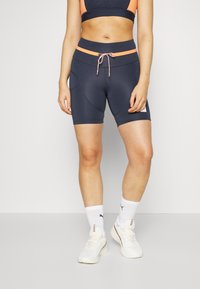 Marinblå atletiska shorts med ljus orange detaljer, elastisk midja, snörning och subtil textur, kombinerat med vita strumpor och sneakers.