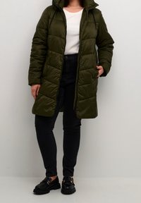 Person in einer olivgrünen Daunenjacke, weißem Hemd, eng anliegenden schwarzen Jeans und schwarzen Slippern, stehend vor einem schlichten hellen Hintergrund.