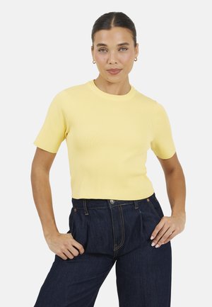 Vrouw met naar achteren gekamd haar, gekleed in een geel topje met korte mouwen en donkerblauwe jeans met hoge taille, poseert met haar handen op haar heupen.