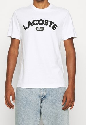Mann trägt ein weißes Lacoste T-Shirt mit schwarzem Logo und hellblauen Jeans, steht vor einem einfachen Hintergrund.