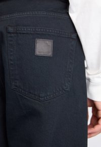 Tamsūs džinsai su užpakaliniu kišeniu, kuriame yra kvadratinis odinis ženklelis su Carhartt logotipu. Medžiaga turi lygų paviršių.
