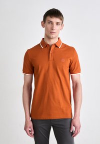 Oranžinė polo marškinė su baltomis detalėmis, su mygtukais užsegama apykakle ir logotipo aplikacija ant krūtinės. Pagaminta iš lygaus audinio, prigludusi konstrukcija.