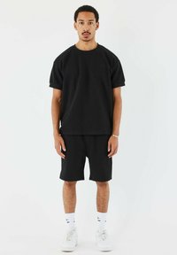 Camiseta negra de algodón con un corte relajado y mangas cortas, combinada con pantalones cortos negros a juego. Zapatillas blancas y calcetines visibles, diseño minimalista.