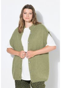 Junge Frau mit welligem Haar trägt eine lockere grüne Strickweste, weißes T-Shirt und grüne Leopardenmuster-Hose, während sie die Knöpfe der Weste befestigt.