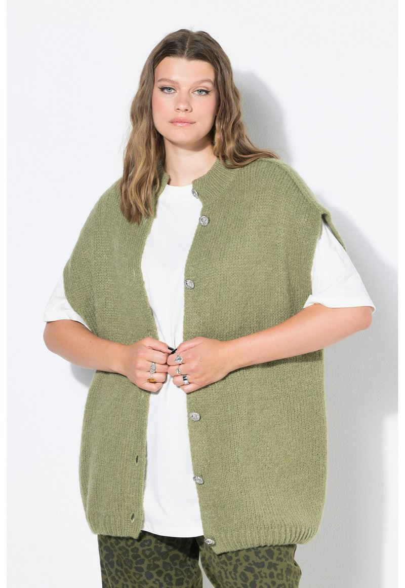 Junge Frau mit welligem Haar trägt eine lockere grüne Strickweste, weißes T-Shirt und grüne Leopardenmuster-Hose, während sie die Knöpfe der Weste befestigt.