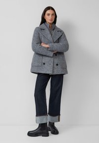 Femme debout, les bras croisés, portant un manteau gris texturé à double boutonnage, un jean foncé à revers et des bottines en cuir noir.
