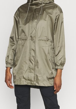 Persona que lleva una chaqueta con capucha de color verde oliva con cordones y puños elásticos, combinada con pantalones negros, de pie frente a un fondo liso.