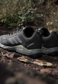Paire de chaussures de randonnée Adidas Terrex noires et grises sur le sol de la forêt avec des feuilles sèches et un feuillage vert en arrière-plan.
