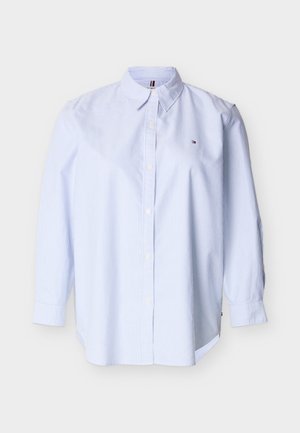 Balta pogājama krekls ar plānām zilām vertikālām svītrām, garām piedurknēm, smailu apkakli un mazu izšūtu logo uz krūtīm uz baltā fona.