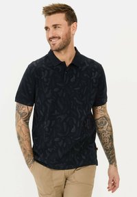 Marineblauw poloshirt met een subtiel bladdessin, korte mouwen en een drieknopen-sluiting. Gemaakt van gladde stof, gecombineerd met beige broeken.