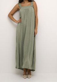 Femme portant une robe maxi sans manches de couleur vert clair avec des points texturés et des bretelles volantes, associée à des ballerines beiges à bout pointu.