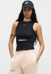 Γυναίκα που φοράει μαύρο καπέλο Nike και μαύρο cropped τοπ χωρίς μανίκια με άνοιγμα, μπεζ φόρμα και φανερό τατουάζ στο χέρι σε λευκό φόντο.
