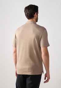 Beige polo-paita lyhyillä hihoilla ja suoran helman kanssa. Valmistettu pehmeästä kankaasta, jossa on kaulus ja takatuuletus. Paita on yhdistetty tummaan housuun.