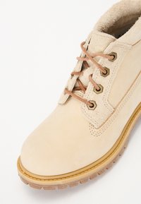 Beige suède enkellaars met bruine veters, metallic rondels en een gestructureerde rubberen zool. Voorzien van verstevigde stiksels en een zachte voering.