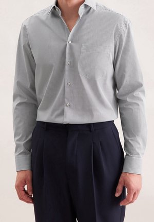 Homme portant une chemise boutonnée à motif gris clair rentrée dans un pantalon plissé taille haute bleu marine foncé, les mains détendues le long du corps.