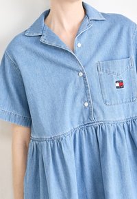 Ljusblå denimklänning med korta ärmar, knäppning framtill och en rynkad midja. Har en bröstficka med en logotagg.