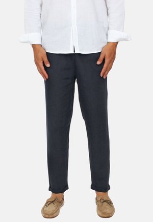 Uomo con pantaloni blu navy, camicia bianca con bottoni e maniche arrotolate, e mocassini color marrone chiaro, in piedi su sfondo bianco.