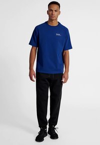 Blå bomullst-shirt med korta ärmar, rund halsringning och en liten logotyp på bröstet, tillsammans med svarta joggerbyxor och mörka sneakers.