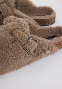 Brown fuzzy slip-on sandals with adjustable metal buckle and cork footbed on a white surface.