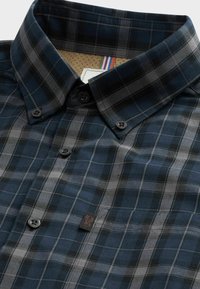 Chemise à carreaux bleu foncé et gris avec boutonnière, col boutonné et petite étiquette noire cousue près de la poche poitrine.