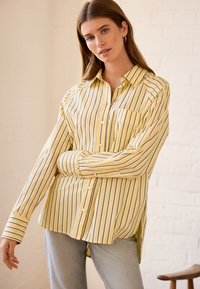 Camicia a righe gialle e bianche con taschino frontale, maniche lunghe e vestibilità rilassata. Abbinata a jeans di jeans chiari.
