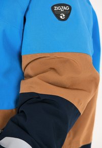 Blå, brun och marinblå färgblockerad jacka med en slät textur och en svart "ZIGZAG" logopatch på överärmen.