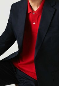 Marinblå kavaj över en röd pikétröja med texturerad yta, med tre knappar och krage, visad i bröst- och axelhöjd.