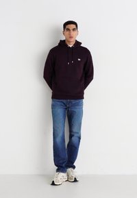 Uomo in piedi contro un muro bianco, indossa un cappuccio nero, jeans blu e sneakers bianche, mani dietro la schiena, espressione neutra.