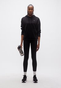 Γυναίκα με μαύρη φούτερ με κουκούλα και κολάν που κρατάει ένα διάφανο μπουκάλι νερού, στέκεται μπροστά σε έναν απλό λευκό φόντο.