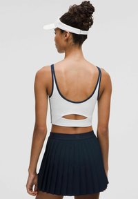 Bijeli atletski crop top s mornarsko plavim akcentima i izrezom, uparen s mornarsko plavom plisiranom suknjom. Uključuje bijeli vizir kao dodatak.